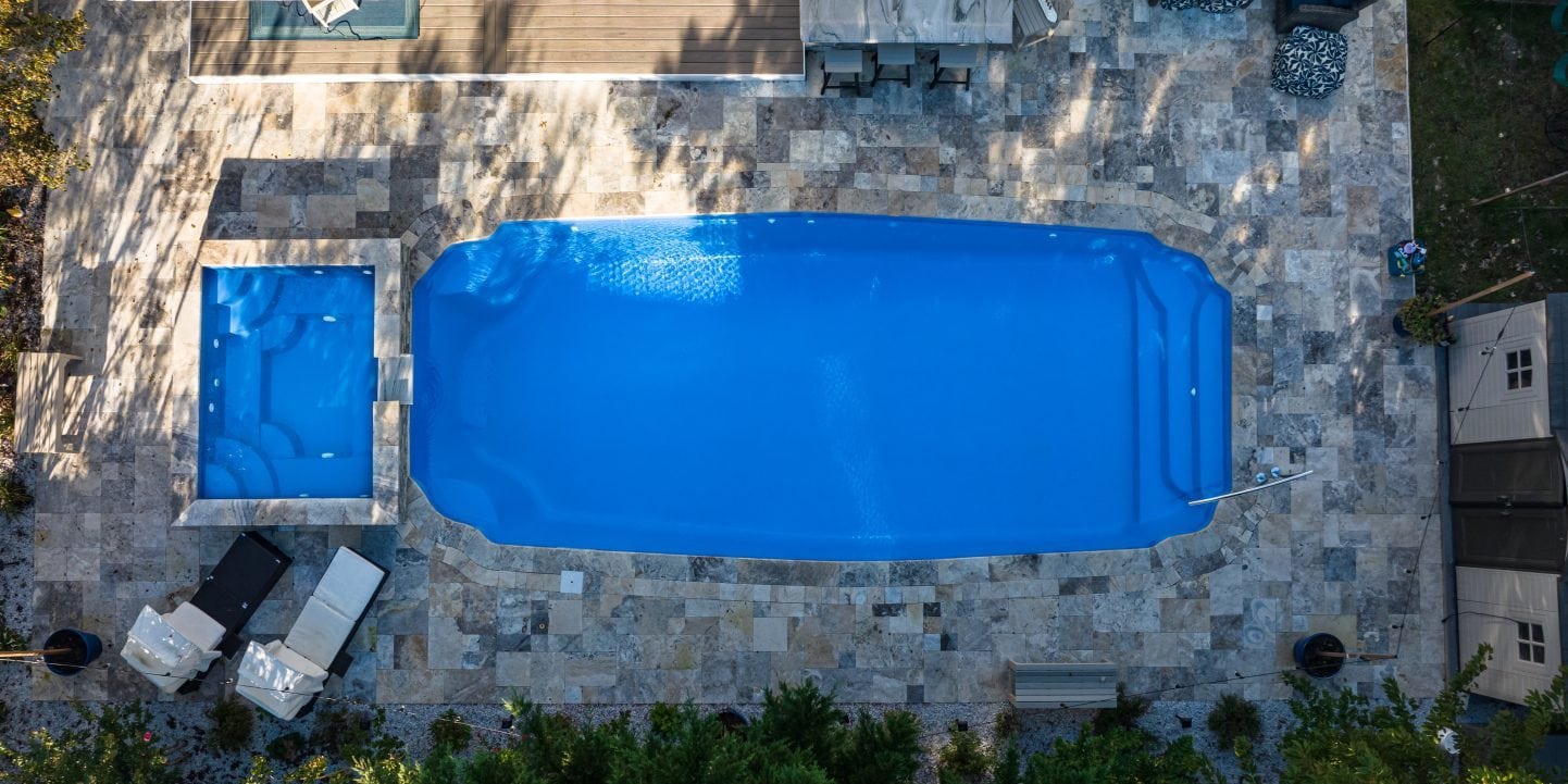15x34-fiberglass-pool-maui-fiberglass-pool-install-6 - MGK Pools Inc Aerial view of a backyard with a large blue rectangular in-ground pool and a smaller square spa surrounded by a stone patio.